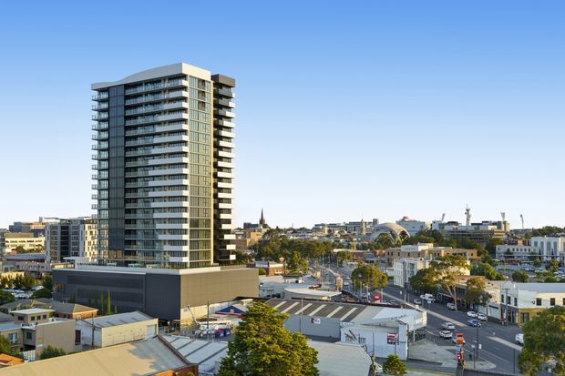 Exterior cityscape view featuring a tall contemporary multi-storey apartment building with extensive balconies and glazed facades, set within a dense urban commercial and residential neighbourhood. Low-rise buildings, streets, trees and a clear blue sky surround the tower.