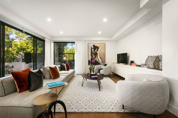 A bright contemporary living room with a large L-shaped light-grey sectional sofa, textured neutral rug, round coffee tables, sculptural armchair, wall-mounted television and long white built-in media cabinetry. Floor-to-ceiling glazing opens to a balcony or exterior garden view, while recessed ceiling lights and timber flooring create a warm, airy setting.