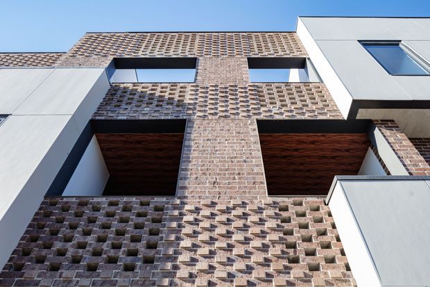 Upward-looking architectural exterior view of a contemporary multi-storey building. The facade combines patterned exposed brickwork with smooth white panelled sections, deep recessed openings, dark timber-clad soffits, and angular projecting forms. Strong sunlight creates pronounced shadows across the textured brick facade against a clear blue sky.