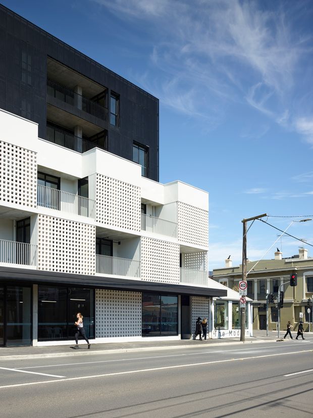 Street-facing view of a contemporary multi-storey mixed-use building with a dark upper facade, white projecting balconies, perforated breeze-block screens, metal balustrades, and large glazed ground-floor commercial frontage. The building occupies a prominent urban intersection beside an older heritage-style streetscape, with tram infrastructure and pedestrians visible.