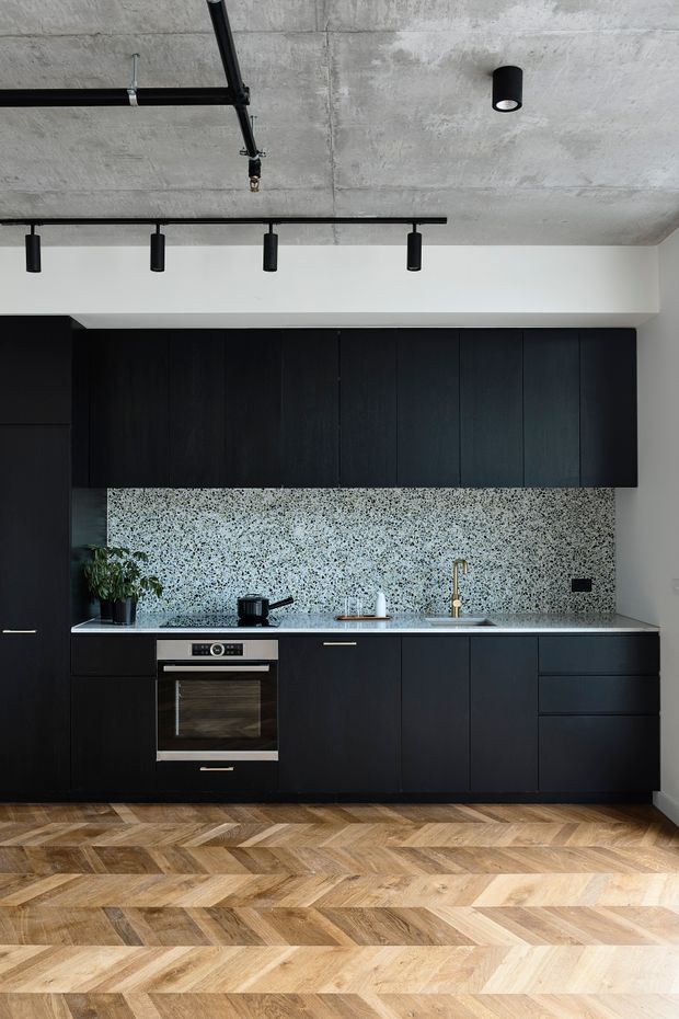 A compact contemporary kitchen with matte black floor-to-ceiling cabinetry, integrated oven, pale stone benchtop, brass tapware, and a terrazzo-style speckled backsplash. The kitchen is set against exposed concrete ceilings with black track and surface-mounted cylindrical lighting, while a warm timber herringbone floor adds contrast.