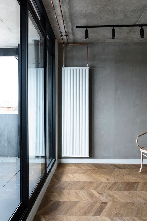 A compact contemporary interior with raw grey concrete walls and ceiling, exposed copper heating pipes, black-framed floor-to-ceiling glazing, and a white vertical-panel radiator. The floor is finished in warm natural timber arranged in a chevron or herringbone pattern. Minimal black track lighting and part of a light wood chair are visible, with a balcony or exterior space beyond the glazing.