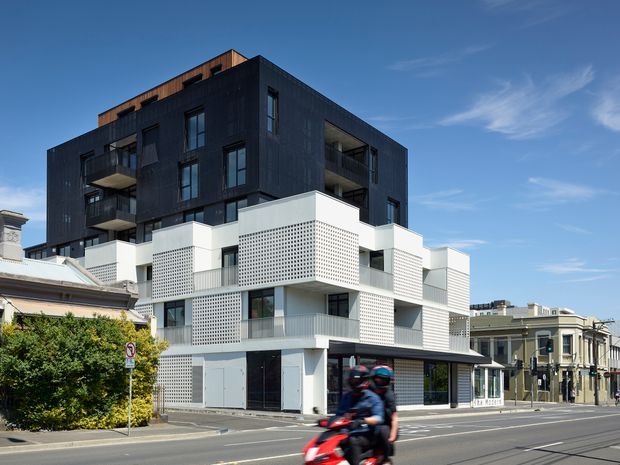 Street-level exterior view of a contemporary multi-storey mixed-use or apartment building. The building combines a dark upper volume with balconies and vertical cladding, and a white lower volume featuring perforated masonry screens, recessed balconies, and ground-floor glazing. It sits prominently on an urban corner street beside older neighbouring buildings.