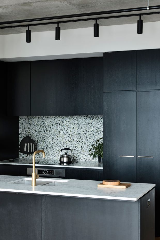 A contemporary dark-toned kitchen featuring full-height charcoal-black cabinetry, a large pale terrazzo-style splashback and island benchtop, integrated appliances, and a prominent brass-gold gooseneck tap. Black cylindrical track lighting is mounted beneath a raw concrete ceiling, creating a refined industrial atmosphere.