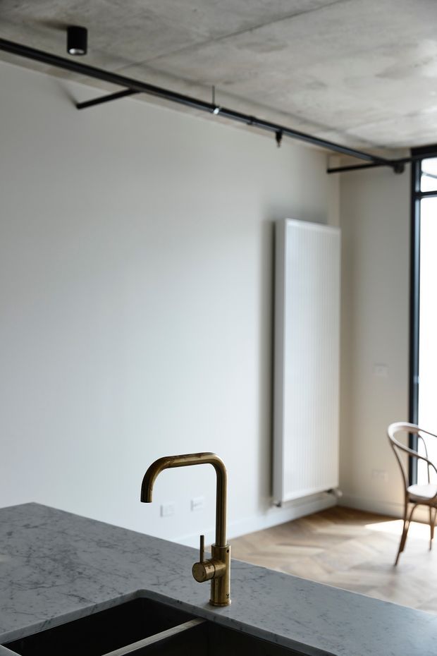 A minimalist kitchen interior focused on a stone island benchtop with an undermount black sink and brass gooseneck tap. The background shows white walls, a tall white panel radiator, herringbone timber flooring, black-framed glazing, and an exposed concrete ceiling with black surface-mounted track lighting. A bentwood-style chair is visible near the window.