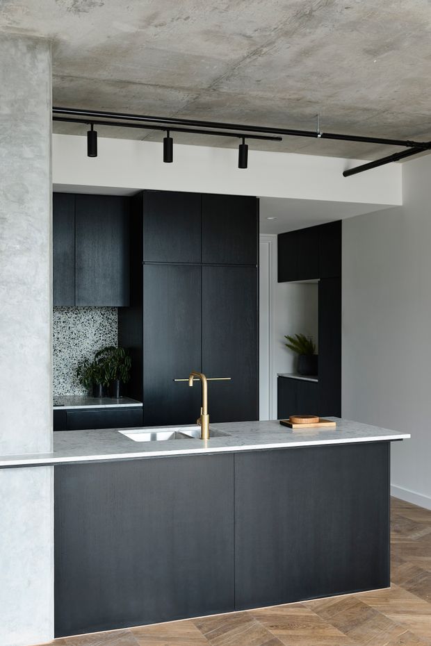 A contemporary kitchen viewed from the living area, featuring a large dark timber island with a pale stone benchtop, integrated double sink and brass gooseneck tap. Matching dark cabinetry extends along the rear wall and into a tall storage bank. A terrazzo-style splashback, raw concrete ceiling and column, black track lighting, white walls, and warm herringbone timber flooring create a refined industrial-modern aesthetic.