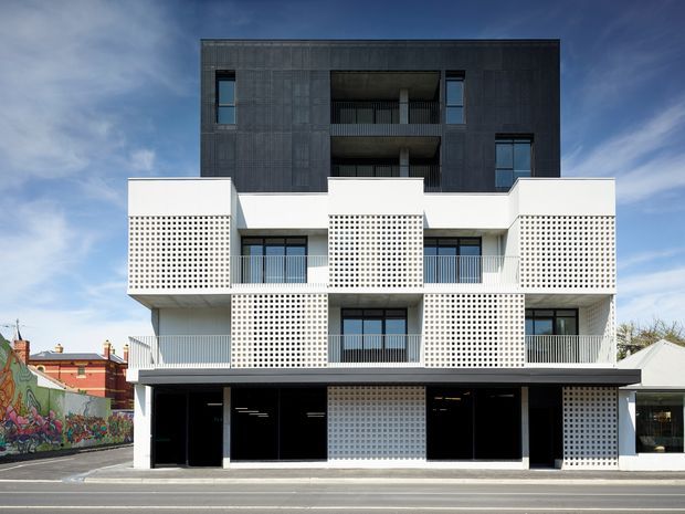 Street-facing multi-storey contemporary apartment building with a strongly geometric facade. The lower and middle levels use white perforated masonry screens around balconies, while the upper level is clad in dark grey textured panels with recessed balconies. Black-framed glazing, a broad dark canopy, and ground-floor commercial or lobby openings face the street. A colourful mural and neighbouring buildings are visible at the edges.