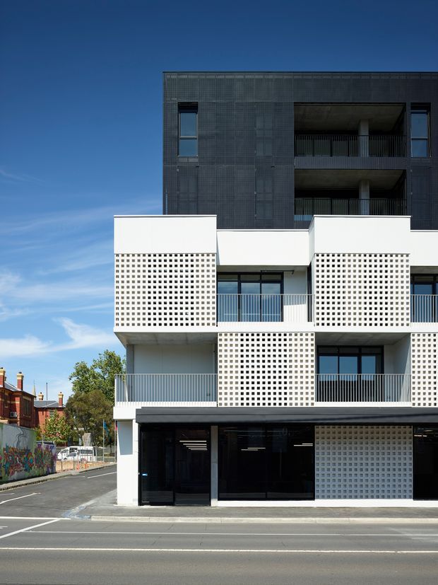 Street-level exterior view of a contemporary multi-storey apartment building. The facade combines dark textured cladding on the upper levels with white rendered sections, projecting balconies, perforated breeze-block screens, glazed ground-floor frontage, and metal balcony railings. Urban streetscape and neighbouring heritage-style houses are visible around the building.