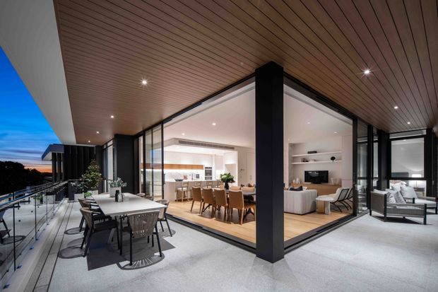 A contemporary multi-level outdoor terrace at dusk, framed by expansive black-framed floor-to-ceiling glazing and a warm timber-lined soffit. The terrace includes an outdoor dining table and chairs, with a covered lounge area visible to the right. Through the glazing, a bright open-plan dining and living interior is clearly visible, featuring a dining table, kitchen, sofa, and built-in shelving. Glass balustrades and dark structural columns reinforce the modern indoor-outdoor connection.