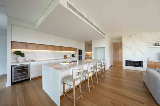 Bright open-plan contemporary kitchen and living area with a large white waterfall island, four bar stools, extensive white cabinetry, a timber-accented storage band, and a veined marble-look splashback. A linear concealed rangehood is integrated into the ceiling, while a fireplace clad in matching veined stone separates the kitchen from the living room. Warm timber flooring and neutral furnishings create an airy, refined interior.