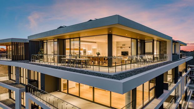 Contemporary multi-storey building exterior photographed at dusk, featuring a prominent flat-roofed upper level with extensive floor-to-ceiling glazing, dark framed windows, projecting balconies, glass balustrades, and warm illuminated interiors. A rooftop terrace with outdoor dining furniture and gravel-edged planting zones is visible, with adjoining units extending into the background.