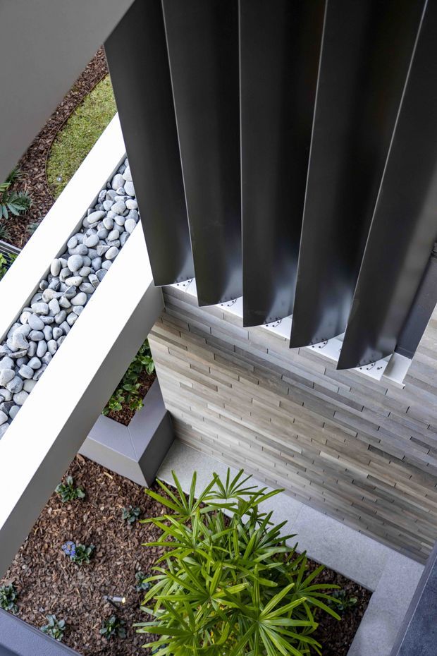 A close-up architectural exterior view looking down onto a landscaped courtyard or entry area. Dark vertical metal fins form a prominent sun-shading or screening element above a pale horizontally coursed masonry wall, with light concrete edging, a white pebble drainage strip, and planted garden beds below.