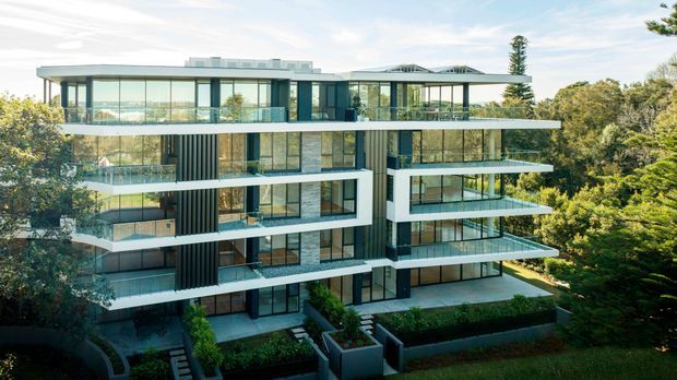 Exterior view of a contemporary multi-storey apartment building set among mature trees. The building features a strongly horizontal composition, extensive floor-to-ceiling glazing, broad projecting balconies with transparent balustrades, dark vertical facade panels, pale rendered or concrete surfaces, and a flat roof with a shallow projecting canopy. Landscaped garden areas surround the lower level.