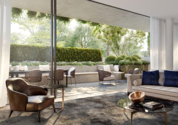A refined contemporary living room opening onto a landscaped terrace through expansive floor-to-ceiling glazing. The room features a cream sofa with deep navy cushions, sculptural brown leather lounge chairs, a dark textured rug, glass and metal coffee table, and a compact dining area. The terrace includes built-in seating, clipped hedges, raised planters, and rounded topiary, creating a calm indoor-outdoor setting.