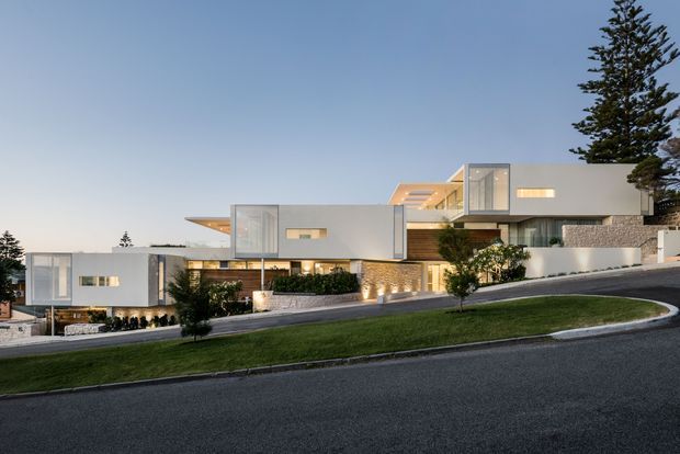 A large contemporary multi-level residence viewed from a sloping suburban street at dusk. The building features crisp white rendered volumes, extensive glazing, projecting flat roof planes, warm recessed lighting, stone-clad lower walls, landscaped terraces, and a manicured grass verge. Tall evergreen trees frame the property, while the stepped massing follows the hillside.