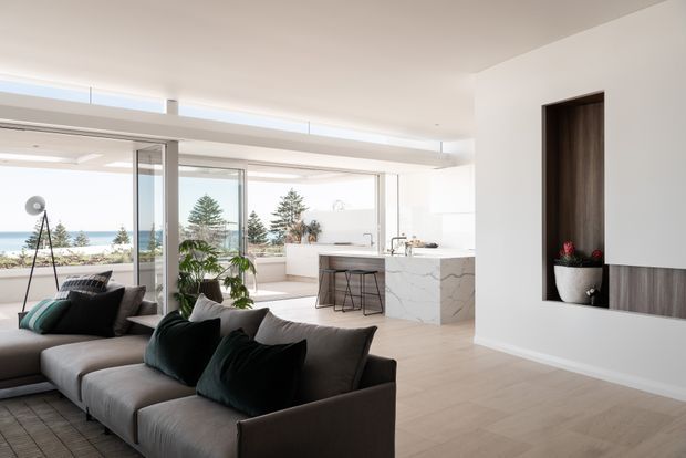 Bright open-plan contemporary living space with a large grey sectional sofa in the foreground, opening onto a minimalist white kitchen with a marble-look island. Full-height glazing provides coastal views and abundant natural light. The room features pale timber flooring, white walls and ceiling, dark timber wall joinery with a recessed display niche, and restrained dark green and black furnishings.