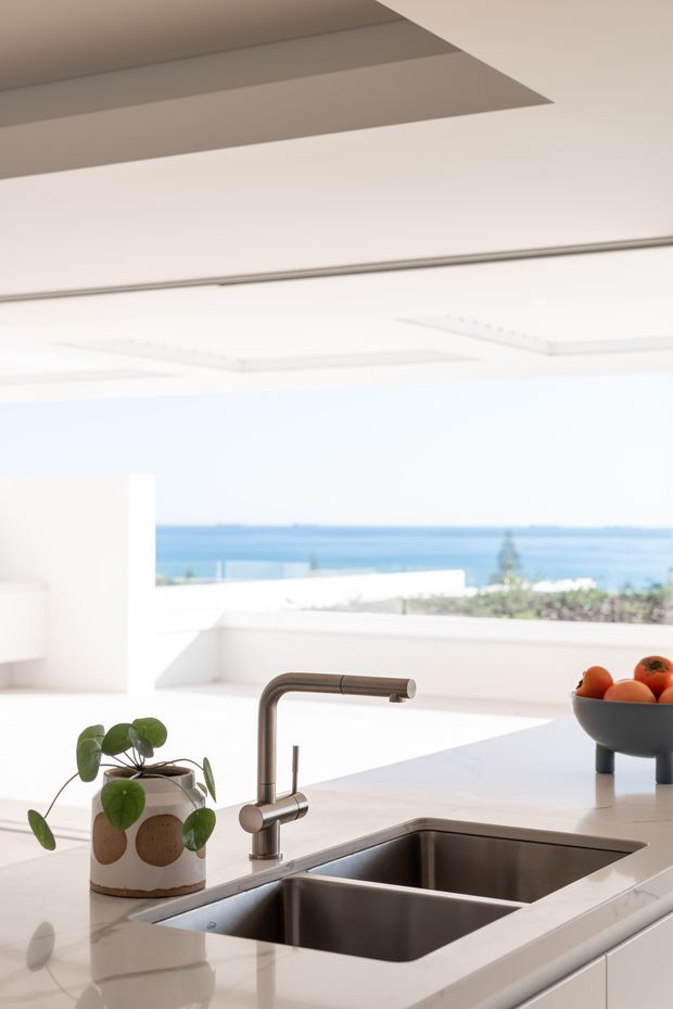 Bright contemporary kitchen interior focused on a large white island with an integrated double stainless-steel sink and curved brushed-metal tap. White cabinetry and surfaces create a clean, minimal setting, with a wide opening or glazing revealing a blue coastal water view beyond. A small potted plant and fruit bowl provide limited decorative accents.