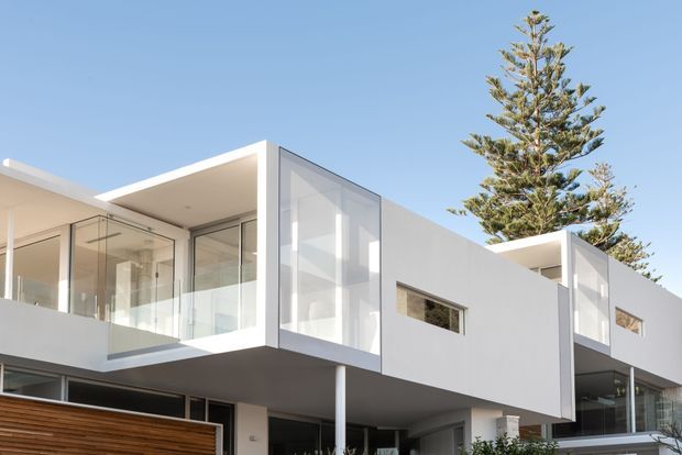 Contemporary multi-level residential facade with crisp white geometric volumes, extensive floor-to-ceiling glazing, projecting upper-storey forms, and a partially visible timber garage or entry door. A tall conifer stands behind the building against a clear blue sky.