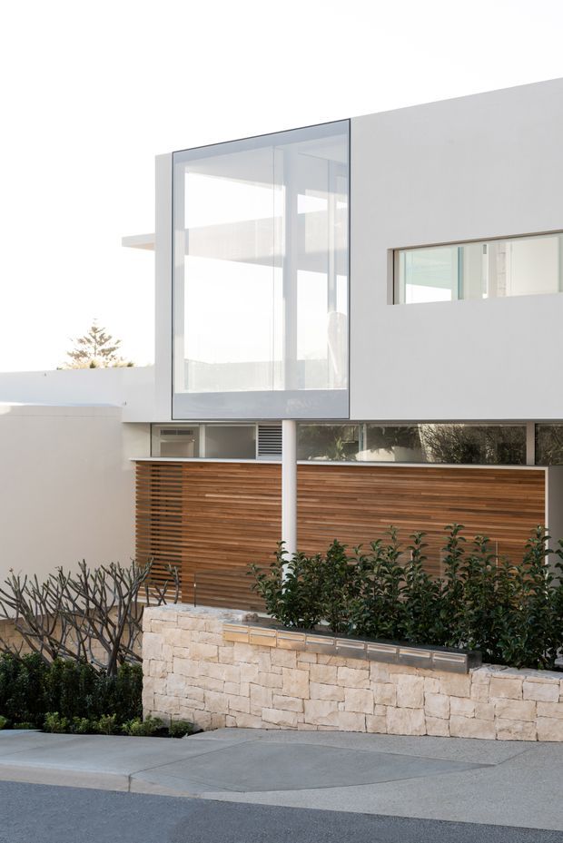 Exterior view of a clean-lined multi-storey residence with white rendered walls, extensive glazed corner openings, narrow horizontal windows, and a warm timber slatted privacy screen at ground level. A pale natural-stone retaining wall, landscaped planting, concrete footpath, and street edge frame the facade. The composition is bright and minimalist with strong horizontal and vertical geometry.