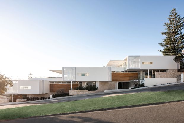 Wide exterior view of a contemporary multi-level residence on a sloping site, composed of crisp white rectangular volumes, extensive glazing, timber screening, and pale stone masonry. The home is set behind a landscaped grass verge and driveway, with a tall evergreen tree at the right edge and clear sky above.