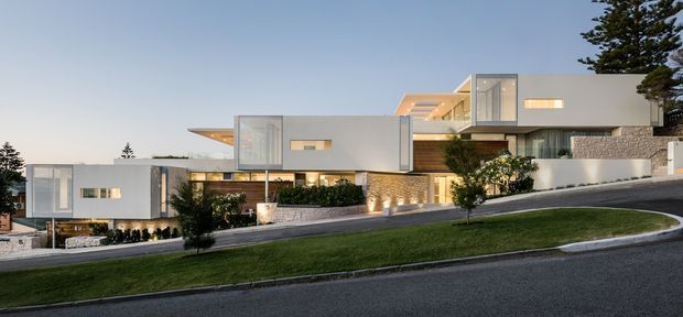 Wide exterior view of a large contemporary multi-level residence on a sloping suburban site. The home features crisp white rendered cubic forms, extensive floor-to-ceiling glazing, projecting cantilevered volumes, warm timber-toned cladding, and textured stone masonry walls. A paved driveway and landscaped lawn occupy the foreground, with mature trees framing the property.