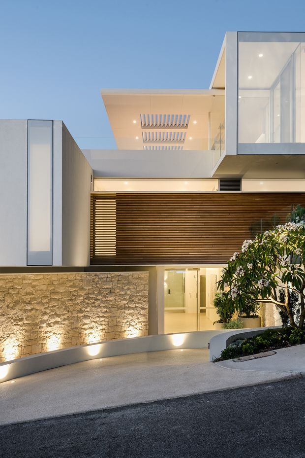A dusk exterior view of a contemporary multi-level residence with crisp white geometric forms, extensive glazing, projecting balconies, timber horizontal screening, and a warm illuminated entry. A textured natural-stone retaining wall and landscaped planting frame the curved driveway and street frontage.