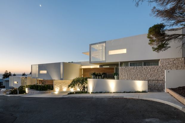 A dusk exterior photograph of a contemporary multi-level residential home on a sloping suburban street. The building features crisp rectilinear volumes, smooth white rendered walls, extensive glazing, warm timber wall cladding, and a prominent textured stone-clad wall. Low landscaped planting, illuminated retaining walls, and a broad driveway frame the facade.