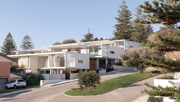 Exterior view of a large contemporary multi-level residence on a sloping suburban site. The home features crisp white rendered volumes, extensive floor-to-ceiling glazing, projecting flat roof planes, balconies, and timber-look wall accents. A curved driveway, landscaped lawns, mature coniferous trees, and neighbouring homes surround the property.