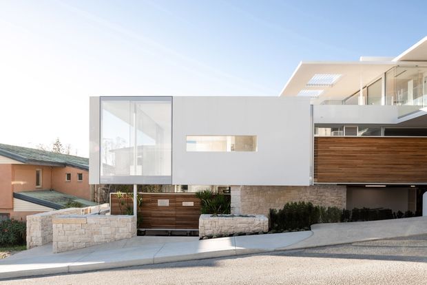 Street-facing view of a contemporary multi-level residence with a clean white rendered facade, extensive glazing, a projecting upper volume, horizontal timber screening, and a sloping driveway. Stone retaining walls, landscaped planting, and a timber garage or entry gate are visible in the foreground.
