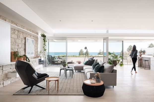 Bright, spacious contemporary open-plan living room with floor-to-ceiling glazing overlooking a coastal landscape. The room features a large neutral sectional sofa, sculptural black lounge chair, layered coffee tables, a textured area rug, indoor plants, and a pale stone-clad feature wall with a fireplace or built-in ledge. A kitchen or bar area is visible at the rear right. The space has a clean, airy, refined interior with strong indoor-outdoor connection.