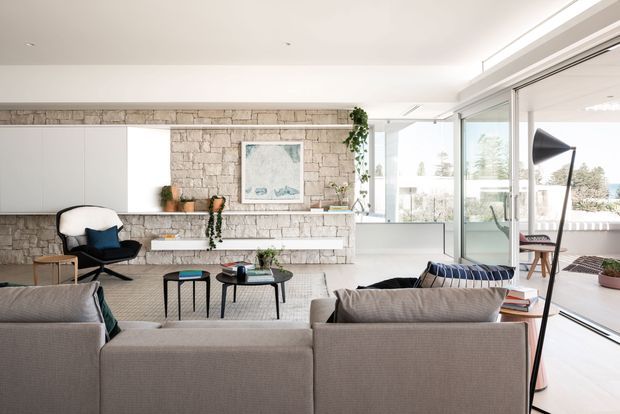 Bright, open-plan contemporary living room with a large neutral sectional sofa in the foreground, mid-century-style lounge chairs and nesting coffee tables, and a stone-clad feature wall with a long white fireplace or media ledge. Extensive floor-to-ceiling glazing opens the room to a light-filled exterior, while built-in white cabinetry and indoor plants complete the refined interior.