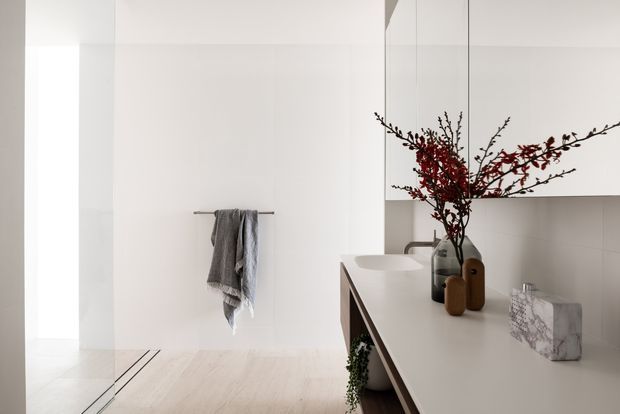 A minimalist contemporary bathroom with a white freestanding basin integrated into a long floating vanity. The room features white wall and ceiling surfaces, pale timber flooring, frameless glass shower glazing, simple wall-mounted cabinetry, and restrained decorative styling with dark vessels and red branch foliage.