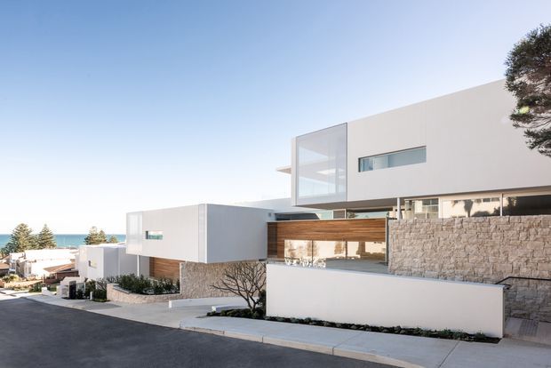 Exterior view of a contemporary multi-level residence on a sloping suburban site. The home features crisp white cubic forms, extensive glazing, warm timber cladding, and a substantial textured stone retaining wall, with glimpses of the sea beyond.