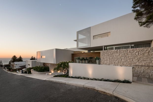 Exterior street-level view of a contemporary multi-level residence on a sloping coastal-looking suburban street. The house features crisp white rendered volumes, expansive glazing, horizontal window bands, a warm timber-lined recessed entry or terrace, and a substantial rough-stone boundary wall. Minimal landscaping and integrated exterior lighting complement the clean geometric composition, with the ocean or distant horizon visible beyond the neighbouring homes.