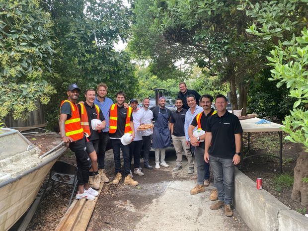 A group of construction and landscaping workers posing together on a narrow outdoor worksite pathway surrounded by dense greenery. Several people wear high-visibility safety vests and work boots, with wheelbarrows, timber planks, concrete edging, tools, and a partially completed garden or construction area visible around them.