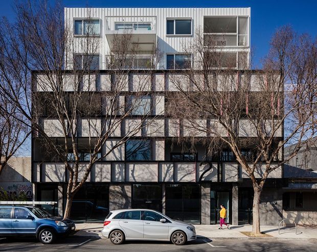 Street-facing view of a contemporary multi-storey apartment or mixed-use building. The facade combines dark ground-level glazing and framing with pale upper-level cladding, projecting balconies, recessed windows, and varied rectangular volumes. Leafless street trees and parked cars occupy the foreground.