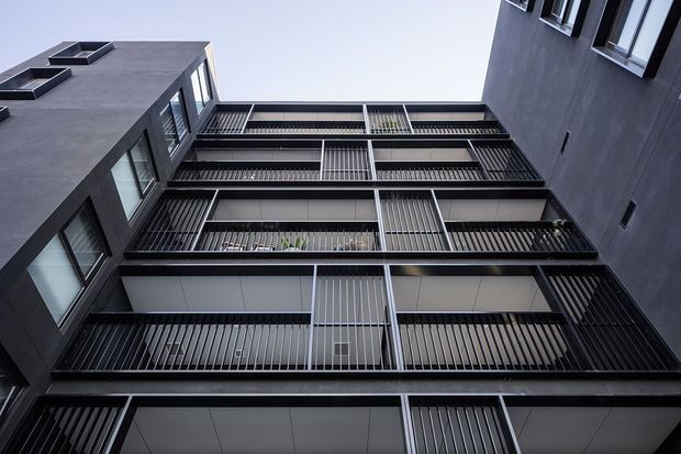 Upward-looking view into a narrow contemporary multi-storey apartment courtyard or atrium. Dark charcoal rendered walls frame stacked balconies with black metal balustrades, vertical screening, recessed glazing, and projecting structural bands. The open sky creates a strong vertical perspective and emphasizes the building’s layered, geometric facade.