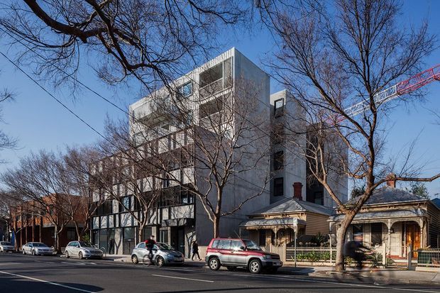 Street-level architectural photograph showing a contemporary multi-storey apartment or mixed-use building with a pale, rectilinear facade, recessed balconies, extensive glazing, and visible rooftop or upper-level forms. It is positioned beside a retained single-storey traditional timber house with a pitched roof, decorative verandah, and ornate frontage. Leafless street trees, parked cars, and an urban streetscape frame the scene.