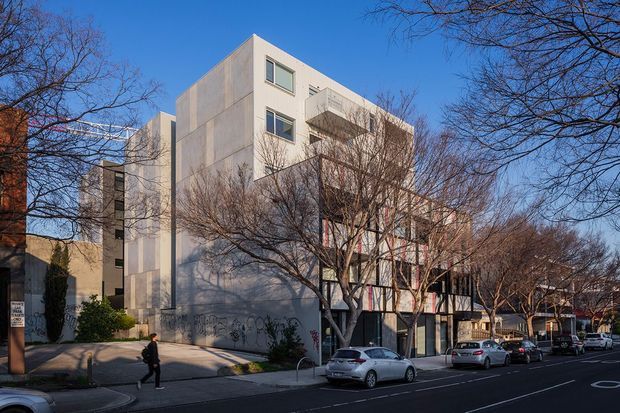 Urban streetscape showing a contemporary multi-storey concrete building with a strongly geometric form, projecting balconies, large windows, and a glazed lower level. Leafless street trees, parked cars, sidewalks, and neighbouring buildings frame the facade.