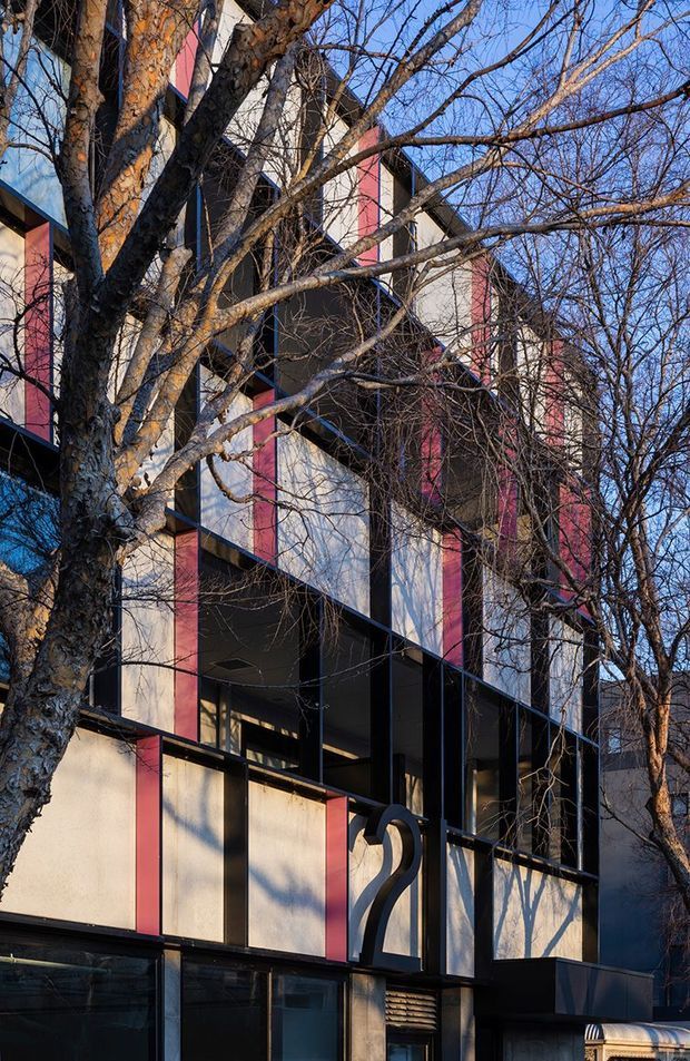 A vertically framed exterior photograph of a multi-storey urban building partially screened by mature leafless trees. The facade features a strong modular composition of dark structural framing, pale wall panels, vivid red vertical accent panels, and extensive glazing. A sculptural curved metal element is visible near the lower entrance area, with a clear blue sky and neighbouring urban context beyond.
