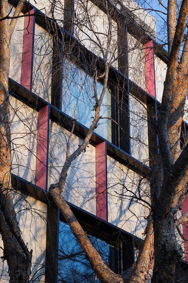 A close-up exterior view of a multi-storey contemporary building partly screened by mature leafless trees. The facade features large reflective blue glazed panels, pale wall panels, dark structural framing, and repeated vivid red vertical accent panels arranged in a strong geometric composition.