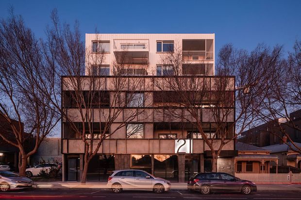 Street-facing view of a contemporary multi-storey residential or mixed-use building with a rectilinear form, layered balconies, dark lower facade, and a pale upper volume. Mature leafless street trees partially screen the building, with parked cars and neighbouring low-rise buildings along the urban street.