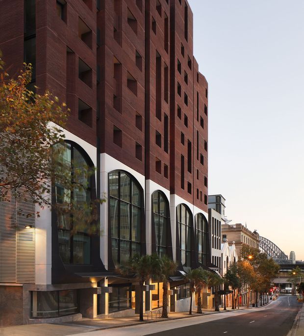 Street-level view of a tall contemporary urban building with a dark red brick facade, repetitive recessed square openings, and a row of very large black-framed arched windows at the lower levels. The building fronts a paved city street with mature street trees and adjacent historic-looking buildings; a large steel bridge is visible in the distance. The architecture combines heavy masonry, strong vertical and horizontal grid lines, and a refined commercial or mixed-use frontage.