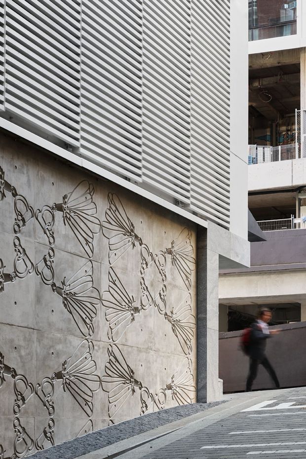 Urban architectural exterior showing a tall concrete wall decorated with a repeated line-art butterfly mural, beneath a large white horizontal louvred screen. Adjacent multi-level concrete construction with open balconies and railings is visible, along with a sloping paved pedestrian ramp and a blurred passerby. The image has a contemporary, industrial character and a monochromatic grey-and-white palette.