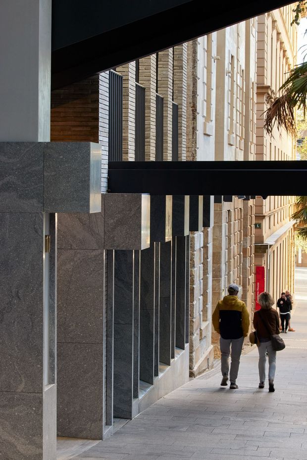 Street-level view of a contemporary urban building facade along a paved pedestrian walkway. The architecture features dark stone panel cladding, projecting vertical fins, black horizontal structural bands, pale textured masonry, and deep-set glazing or openings. An adjacent historic-looking stone facade and pedestrians are visible, with palm foliage at the far right.