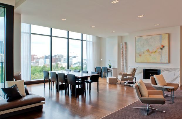 Bright, spacious contemporary open-plan living and dining area with floor-to-ceiling windows overlooking an urban landscape. The room features dark timber flooring, a large dark dining table with upholstered chairs, a lounge seating area, a marble fireplace surround, recessed ceiling lighting, and a large abstract artwork. The palette is neutral and refined with abundant natural light.
