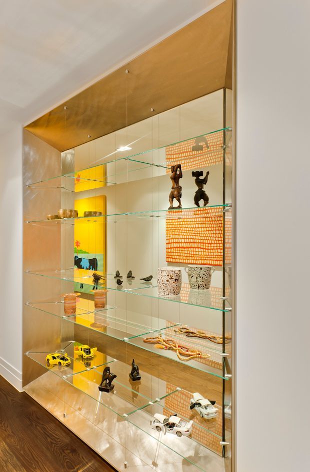 A tall built-in display cabinet with mirrored backing, clear glass shelves, and warm wood-toned paneling. The shelving showcases sculptures, pottery, jewellery or beads, and small decorative objects, with bright artwork reflected in the mirror. The cabinet is set into a neutral interior beside dark timber flooring.