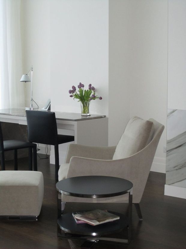 A refined, minimalist sitting and work area with a cream upholstered lounge chair and ottoman, a black round two-tier side table, and a compact desk with black chairs. The room features white walls, dark timber flooring, a marble-clad vertical feature at the right, and soft daylight through sheer curtains. A small vase of purple flowers and a slender desk lamp provide subtle accents.