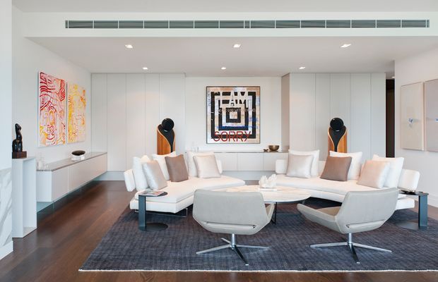 Bright, spacious contemporary living room with a large white modular sectional, sculptural swivel lounge chairs, dark timber flooring, a large charcoal area rug, and extensive floor-to-ceiling white built-in cabinetry. Recessed ceiling lighting, linear air-conditioning vents, modern artwork, and carefully styled shelving create a clean, refined interior.