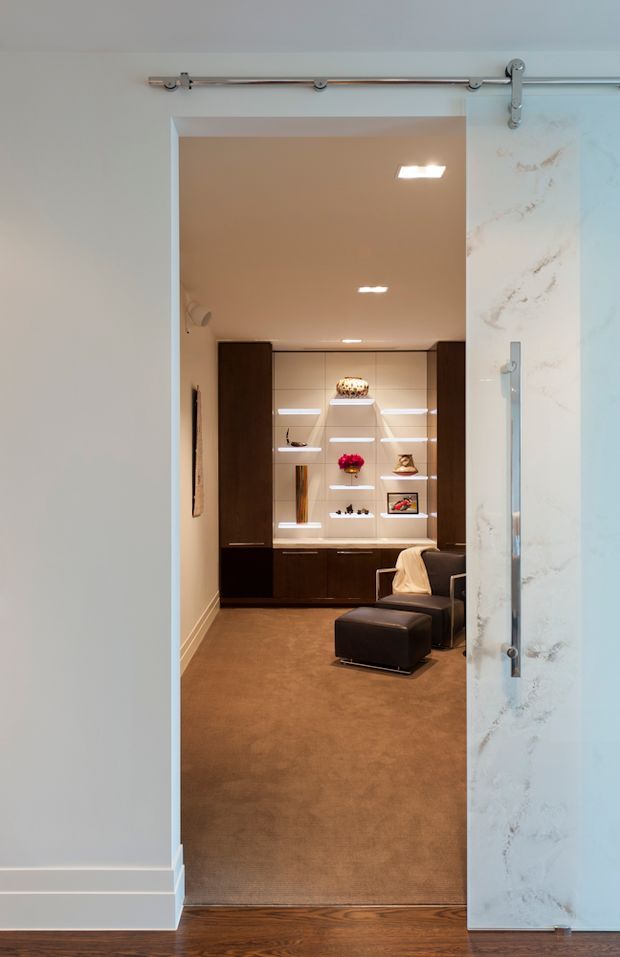 View through a large sliding barn-style glass door into a compact, refined lounge or sitting room. The room has warm beige carpet, dark timber built-in cabinetry with illuminated open display shelves, a dark leather armchair and ottoman, recessed ceiling lighting, and a tall white marble-look door panel and trim. The foreground includes dark timber flooring and white walls.